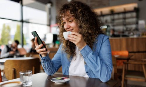 Happy young woman using mobile phone and enjoying coffee at cafe