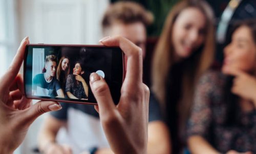 Female hands taking photo with smartphone of young cheerful friends, blurred background.